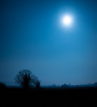 Blue nighttime sky with a silhouetted ground and overexposed moon